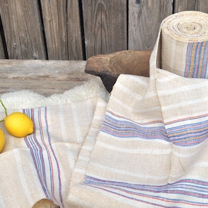 May include: A roll of natural linen fabric with blue, red, and brown stripes. The fabric is rolled up and sitting on a wooden table with two lemons and a sprig of green leaves.