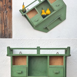 May include: A green wooden wall shelf with three compartments and a small drawer. The shelf is mounted on a white wall and has gourds in the top compartments.