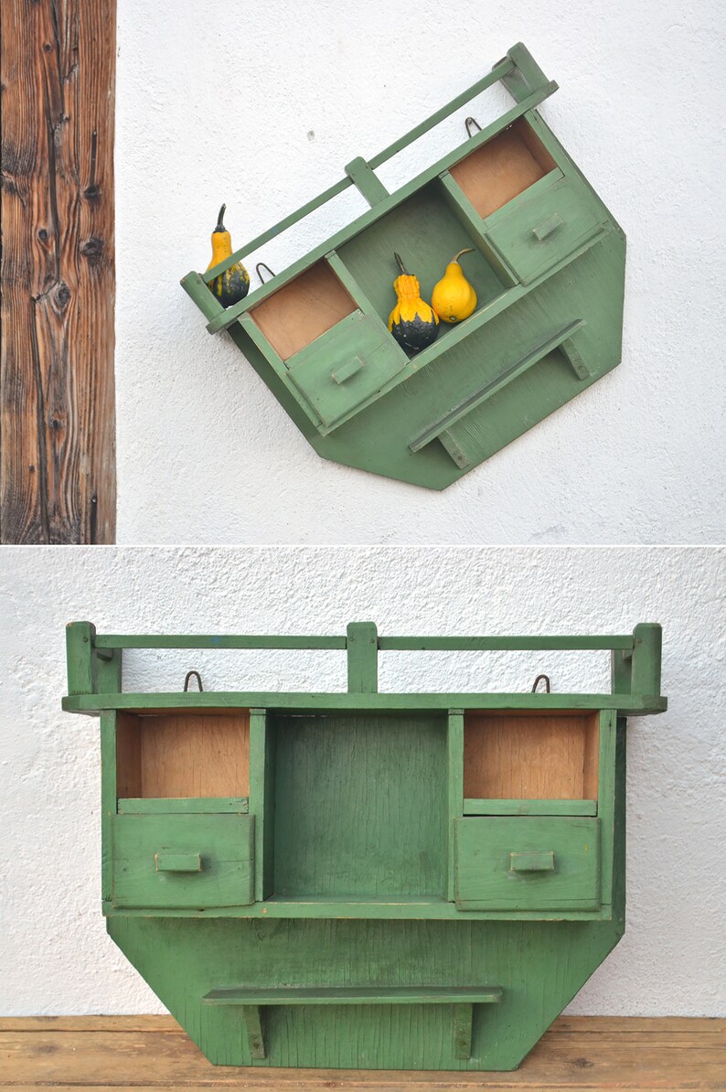 May include: A green wooden wall shelf with three compartments and a small drawer. The shelf is mounted on a white wall and has gourds in the top compartments.