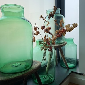 May include: Three green glass jars with a dried flower arrangement in the center jar. The jars are sitting on wooden stools.