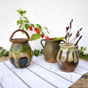 Puede incluir: Tres macetas de cerámica vintage con un esmalte verde, marrón y blanco. Las macetas están dispuestas sobre una superficie rayada blanca y negra. Las macetas están decoradas con una variedad de patrones y diseños.