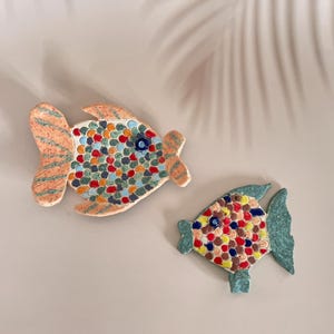 Op de afbeelding: Twee keramische vis wanddecoraties, een groter dan de andere, met een mozaïek van kleurrijke cirkels op hun lichamen. De grotere vis is oranje en wit met groene en blauwe accenten. De kleinere vis is groen met blauwe en rode accenten.