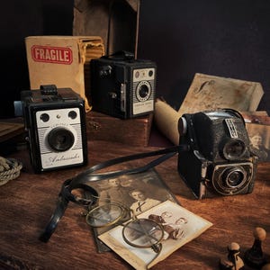 以下が含まれることがあります： 木製の表面に配置された、Coronet Ambassador、Poplar Model、Ful-Vueなどのヴィンテージカメラのコレクション。 カメラは黒とシルバーで、レンズとダイヤルが付いています。 「FRAGILE」と書かれた箱が背景にあります。