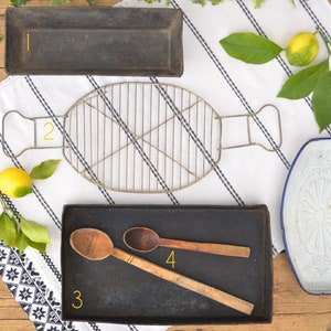 May include: Five vintage kitchen items on a wooden surface. A black metal baking sheet, a wire rack, a wooden spoon, a black metal baking sheet with two wooden spoons, and a white enamelware dish with blue trim.