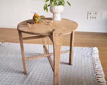 Puede incluir: Una mesa redonda plegable de madera con un diseño tallado único en las patas. La mesa está hecha de madera marrón claro y tiene un acabado natural. Está sentada sobre una alfombra gris y blanca con flecos.
