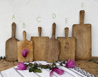 May include: Six antique wooden cutting boards of varying sizes and shapes are displayed against a white wall. The boards have handles and show signs of wear, with a rustic, aged appearance. A white cloth with black stripes and floral accents is in the foreground.