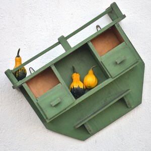 May include: A green wooden shelf with three compartments and a drawer. The shelf is mounted on a white wall and decorated with gourds.