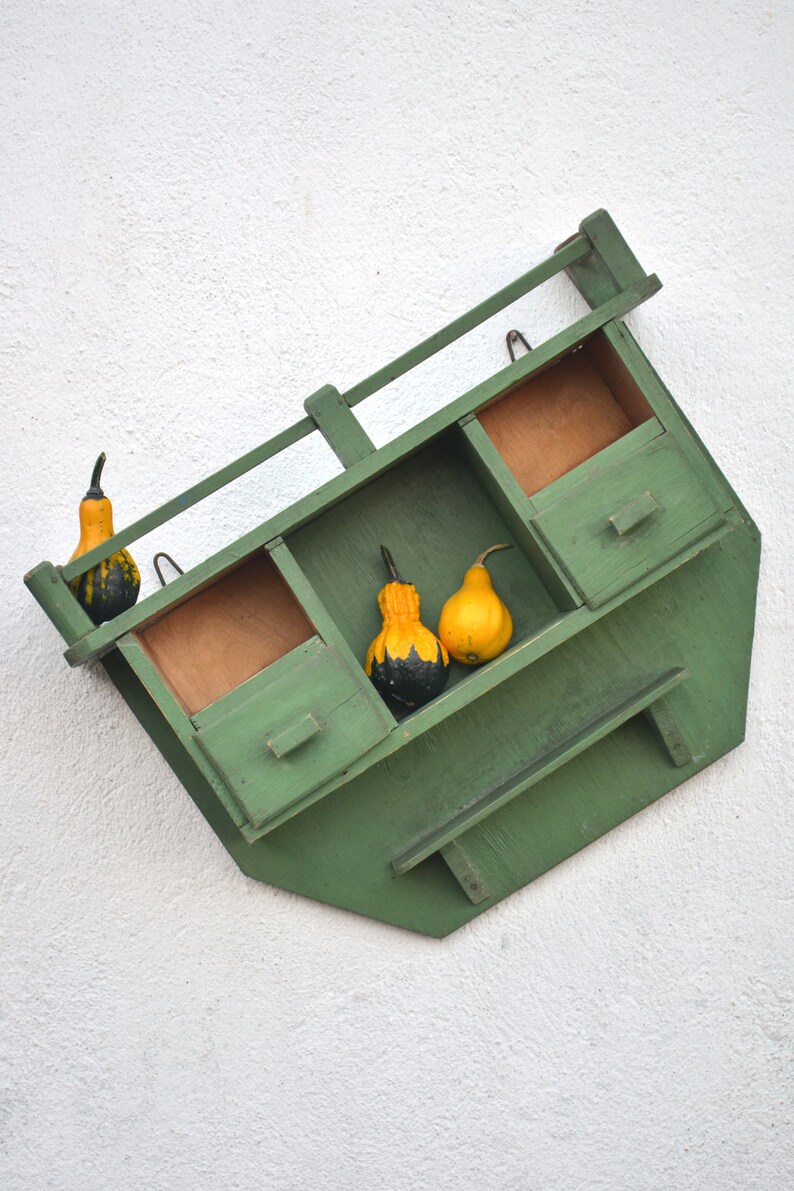 May include: A green wooden shelf with three compartments and a drawer. The shelf is mounted on a white wall and decorated with gourds.