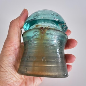 Può includere: Un isolatore elettrico vintage in vetro blu acqua con una parte superiore arrotondata e una base cilindrica. Il vetro presenta una striscia verticale color ruggine e scritte in rilievo che riportano "RD A" con il numero "1" visibile vicino al bordo inferiore.