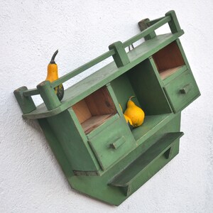 May include: A green wooden wall shelf with two compartments and a small drawer. The shelf has a small ledge on the bottom. There are three gourds on the shelf, two yellow and one green.