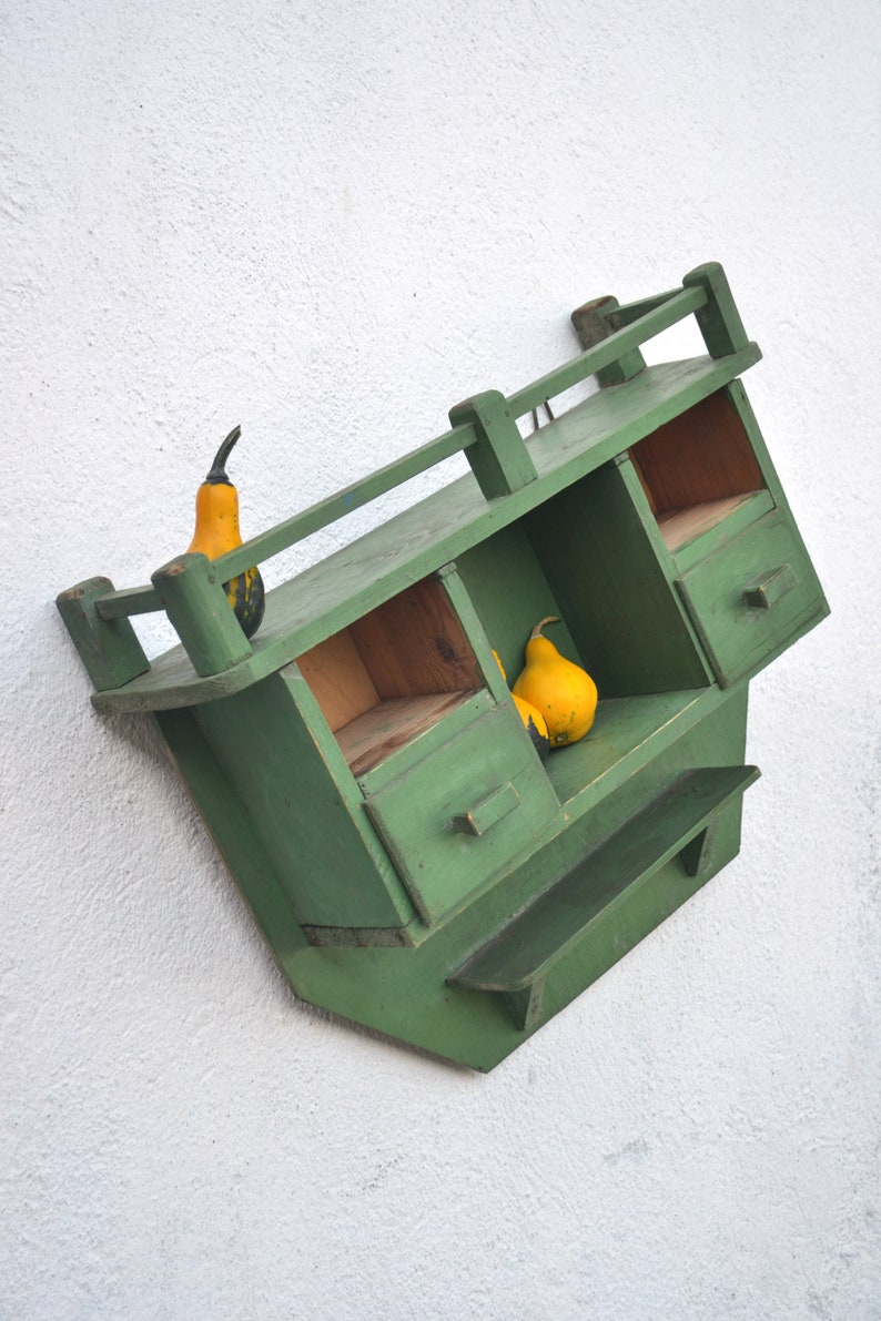 May include: A green wooden wall shelf with two compartments and a small drawer. The shelf has a small ledge on the bottom. There are three gourds on the shelf, two yellow and one green.