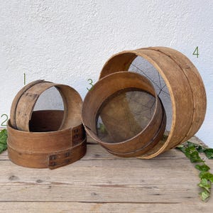 May include: Three antique wooden sieves of varying sizes are arranged on a wooden surface. The sieves are made of light brown wood with metal mesh screens. The largest sieve is partially open, revealing the mesh.