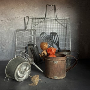 Op de afbeelding: Een stilleven met een verzameling vintage keukengerei, waaronder draadgaas, een garde, een rasp en twee metalen containers. De gereedschappen zijn gerangschikt op een grijs oppervlak met gedroogde bloemen en een paar rode bessen.