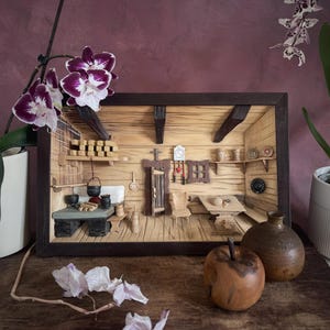 Op de afbeelding: Een houten diorama van een rustiek kamerinterieur met een tafel, een stoel, een wandklok, planken met miniatuur keukengerei, een deur en een kachel. De scène is gevat in een donker rechthoekig frame met wanden en vloer van licht hout.