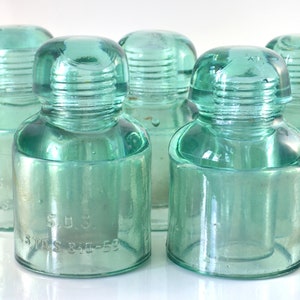 May include: Five vintage green glass insulator bottles with threaded tops. The bottles are clear and have a slight greenish tint. The bottles are all the same size and shape. One bottle has the text "5 U.S.  WIRES 345-52" printed on the side.