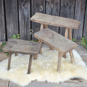 May include: Three rustic wooden stools of varying heights, with a white sheepskin rug underneath. The stools are made of rough-hewn wood and have a natural, weathered finish.