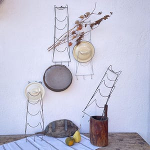 May include: A rustic display featuring metal plate racks, holding vintage plates and lids. The arrangement includes a wooden trough, gourds, and dried branches, all set against a white wall and a wooden table with a striped cloth.