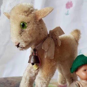 May include: A vintage stuffed lamb with a bell around its neck and a small doll wearing a green hat and burgundy overalls. The lamb is cream-coloured with green eyes and a tan ribbon. The doll is seated next to the lamb.