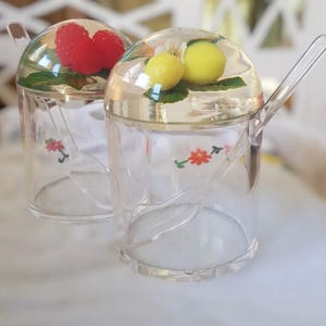 May include: Two clear glass sugar dispensers with spoons. One dispenser has a red berry on top and the other has a yellow lemon on top.