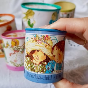 May include: A blue and white metal tin with a floral design and two children on the front. The tin has the text "Monday's child is fair" on the bottom.