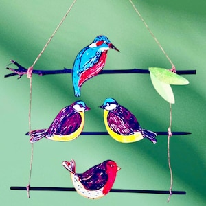May include: A decorative mobile with four colourful bird cutouts and a green leaf. The birds are perched on black horizontal bars and a small branch, suspended by twine against a green backdrop. The birds feature blue, red, yellow, and purple details.