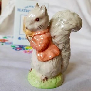 May include: A small ceramic figurine of a grey squirrel wearing a pink jacket and holding a nut. The figurine is standing on a green base and has a detailed face.