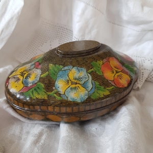 May include: An antique, oval tin box with a hinged lid. The box is decorated with colourful pansies in yellow, blue, and orange, with green leaves against a dark brown background. The lid has a small, raised oval detail.