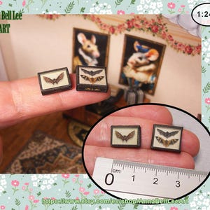 May include: Miniature framed bat art pieces. Each tiny frame displays a detailed bat illustration. The frames are held between fingers, with a ruler showing scale. The background includes miniature paintings and floral wallpaper.
