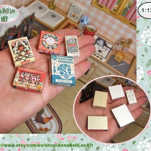 May include: Miniature Christmas ornament boxes in various designs, including a box with the text "Shiny & Brite". The boxes are held in a hand, with a miniature kitchen scene in the background. The text "Anna Bell Lee ART" is visible.
