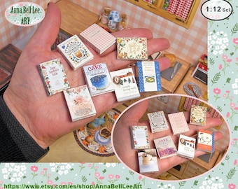 May include: A collection of miniature books with various titles, held in a hand. The books feature floral and patterned covers, with titles like "Cottagecore" and "Cake Decorating." The image includes the text "Anna Bell Lee ART" and "1:12 Scl."