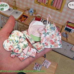 Peut inclure: Accessoires de cuisine miniatures, dont des assiettes, des serviettes et un tablier, sont tenus dans une main. Les articles présentent un motif floral avec des roses roses et vertes. L'arrière-plan montre une scène de cuisine miniature.