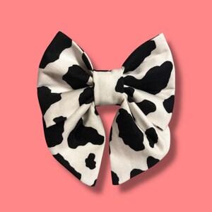 May include: A white and black cow print bow tie. The bow is made of fabric and has a classic bow shape.