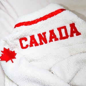 May include: White fleece jacket with red accents and the word "CANADA" in bold red letters. A red maple leaf emblem is on the left sleeve. The jacket has a soft, textured appearance.