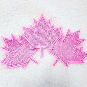 May include: Three pink embroidered maple leaf patches. Each leaf has a detailed, textured design and a bright pink outline. The patches are arranged on a white, textured background.