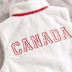 May include: A white sleeveless vest made of soft, fuzzy material. The garment features a high collar with a red trim along the base. The word CANADA is embroidered across the upper back in bold, red, block-style capital letters with a white outline.