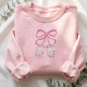 Puede incluir: Sudadera rosa claro con cuello redondo y mangas largas. La parte delantera presenta un diseño bordado de dos latas de refresco Dr. Pepper Diet atadas con un lazo rosa. Las latas tienen un estampado floral rosa.