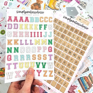 Può includere: Una collezione di adesivi con alfabeto e tessere Scrabble colorate. Gli adesivi dell'alfabeto presentano lettere in vari colori con un bordo cucito. Gli adesivi Scrabble hanno tessere di legno con lettere e valori. Il testo "simplywatercolorco" è visibile.