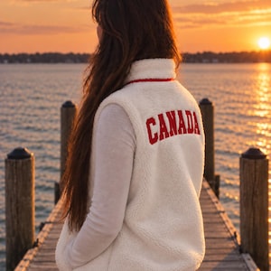 May include: A cream-colored, sleeveless sherpa fleece vest featuring a high collar with red trim. The back of the vest displays the word "CANADA" embroidered in bold, red, capital letters. The garment has a textured, fuzzy exterior finish.