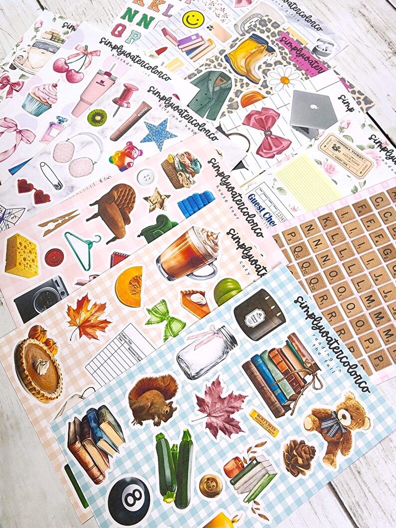 May include: A collection of decorative stickers with various illustrations, including food, books, and seasonal items. The stickers feature a variety of colors and designs, with some including text. The stickers are arranged on a light-colored surface.