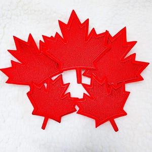 May include: A collection of red maple leaf patches, arranged on a white, textured surface. The leaves are a vibrant red, with detailed stitching outlining their shape. Ideal for crafting or adding a patriotic touch.