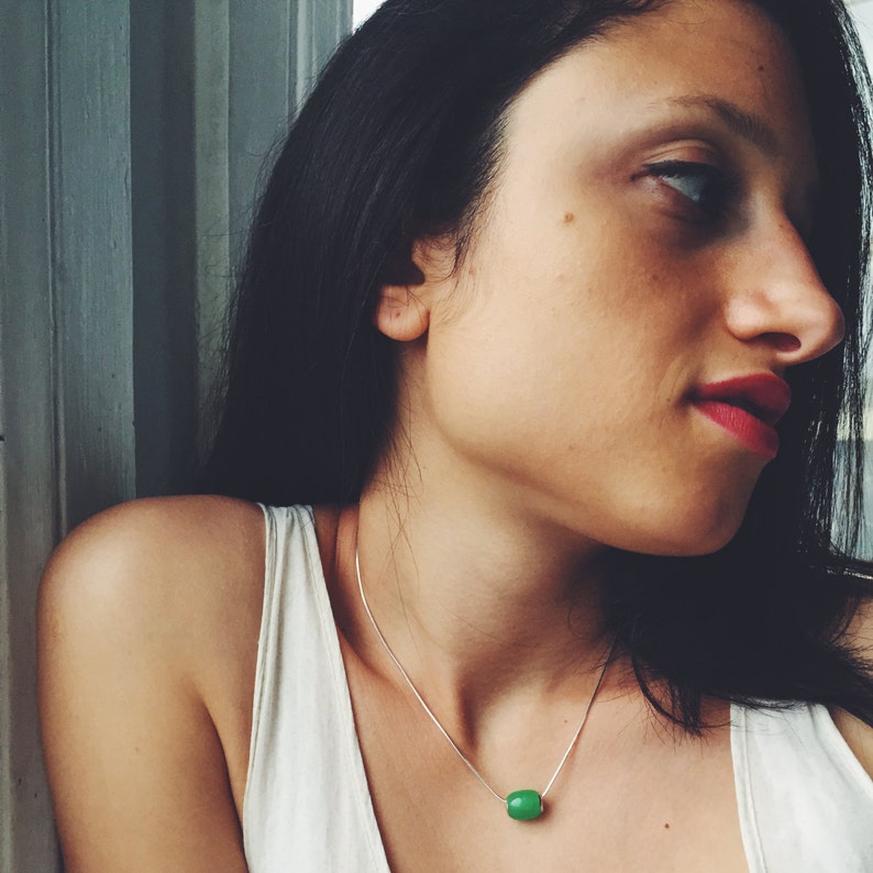 May include: A silver necklace with a small, bright green, cylindrical bead pendant. The necklace is worn by a person wearing a white tank top. The person has dark hair and red lipstick.