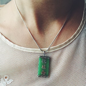 May include: A silver necklace with a rectangular green jade pendant. The pendant has Chinese characters in a vertical arrangement. The necklace is worn over a white, mesh-like top.