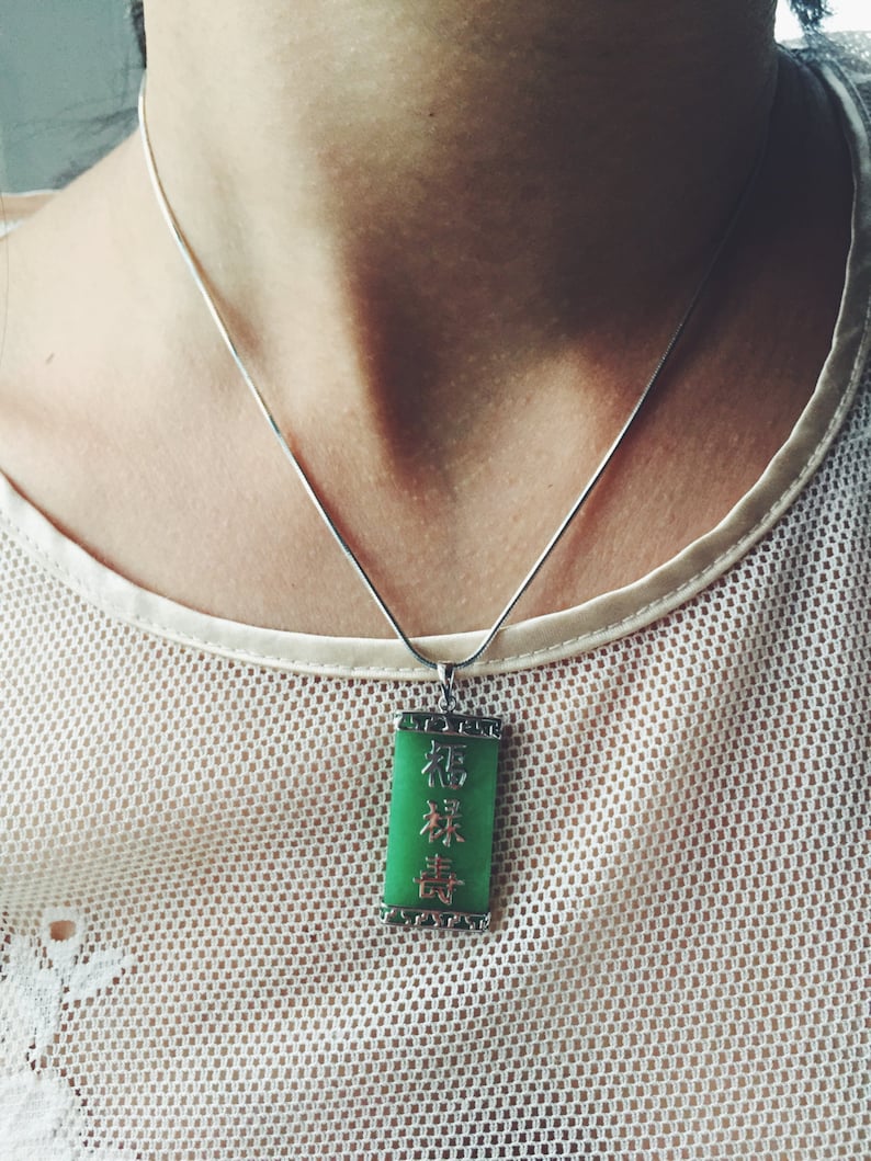 May include: A silver necklace with a rectangular green jade pendant. The pendant has Chinese characters in a vertical arrangement. The necklace is worn over a white, mesh-like top.