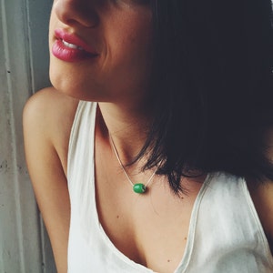 May include: A silver necklace with a bright green, cylindrical bead pendant. The pendant is the focal point, suspended from a delicate chain. The necklace is worn against a white tank top.