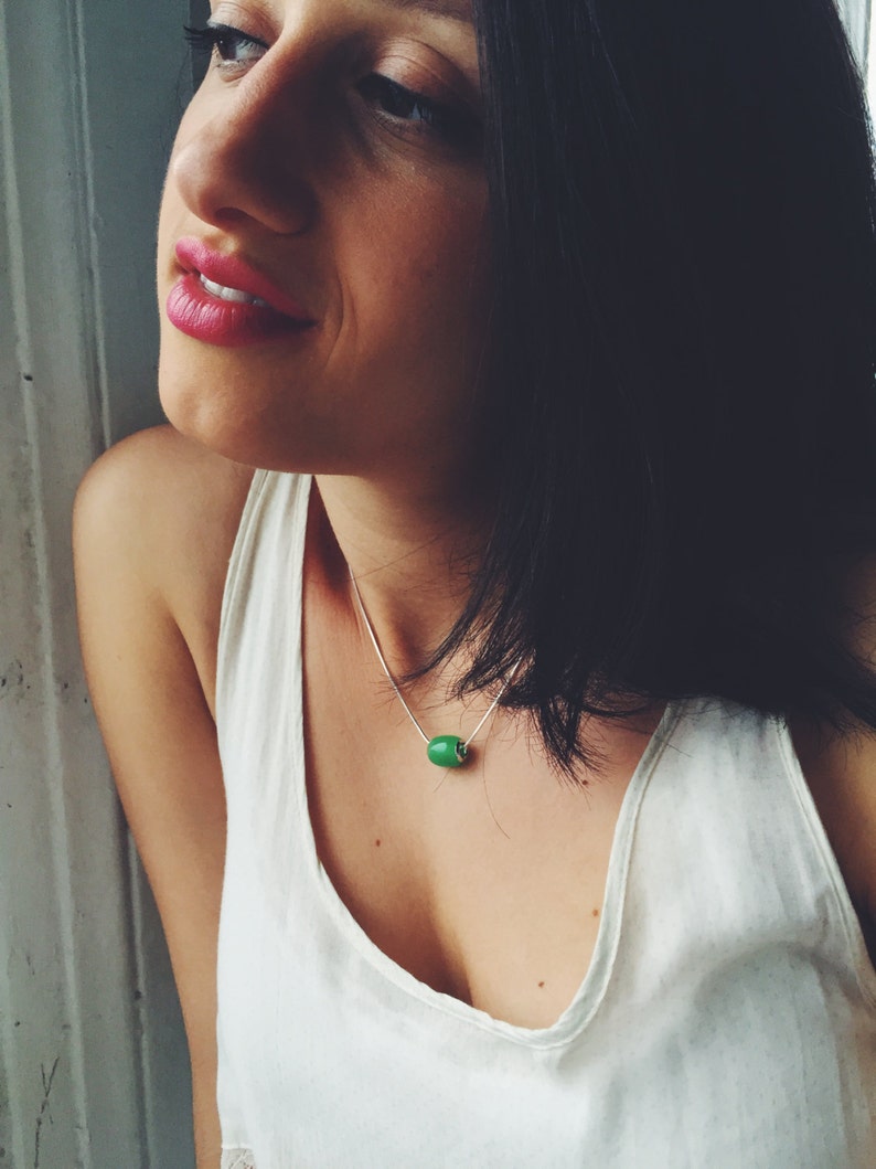 May include: A silver necklace with a bright green, cylindrical bead pendant. The pendant is the focal point, suspended from a delicate chain. The necklace is worn against a white tank top.
