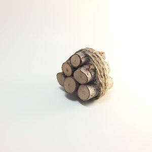 Può includere: Un fascio di piccoli tronchi di legno naturale legati insieme con lo spago. I tronchi sono disposti a forma di piramide e hanno un colore marrone chiaro.