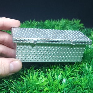 May include: A small, silver, rectangular box with a textured lid. The box is sitting on a bed of green artificial grass.