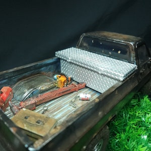 Peut inclure: Un plateau de camionnette noire rouillée avec une boîte à outils en tôle d'aluminium à motif diamant, une tronçonneuse, un cric et d'autres outils. Le plateau du camion est sale et a une surface peinte bleue et blanche.