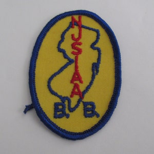 May include: An oval-shaped embroidered patch with a yellow background and a blue border. It features a blue outline of the state of New Jersey, the red text NJSIAA vertically inside the outline, and the blue letters B B at the bottom.