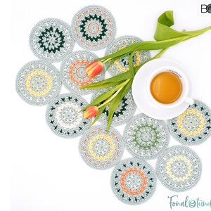 May include: A set of ten crocheted coasters in shades of grey, green, yellow, and orange. The coasters are arranged in a circle with a cup of tea and a bouquet of tulips in the centre. The coasters are handmade and feature a variety of intricate patterns.